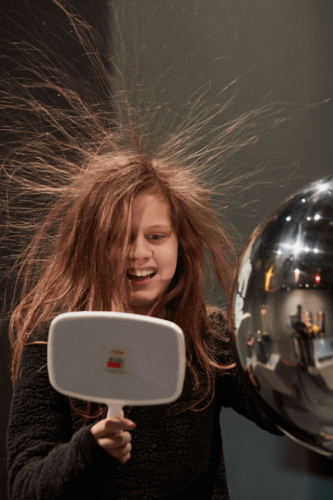 Une petite fille touche d’une main un générateur de Van de Graaff et tient dans l’autre un miroir pour observer ses cheveux relevés en l’air à cause de l’électricité statique généré par l’appareil.
